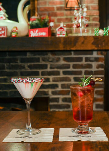 two cocktails on a table in front of the fire. an opaque white drink in a martini glass with red and green sprinkles on the rim. a red drink in a footed highball glass garnished with rosemary